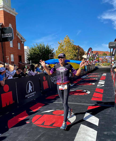 Myron Pozniak, MD, FACR, crosses the finish line at the St. George Ironman 70.3 World Championship, carrying the flag of his native Ukaine